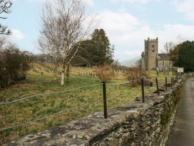 A stone wall with metal fencing beside a path leading to a church with a clock tower and a graveyard with tombstones near trees at Swifts Reach Lodge in Kirkstone 10 Limefitt Park near Windermere