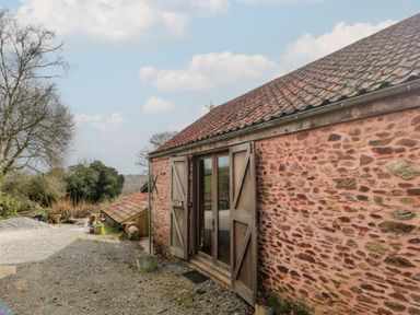 An exterior view of a stone building with wooden doors open on a gravel ground with trees in the background at The Barn in Spaxton
