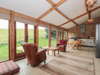 A living area with two leather armchairs a wooden coffee table a dining table with chairs kitchen cabinets and large windows showing greenery outside at The Barn in Spaxton