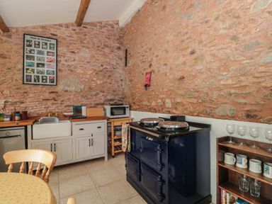 A kitchen with a stone wall, sink, microwave, stove, wooden chairs and shelves with cups and glasses at The Barn in Spaxton