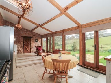 A dining area with a round table and wooden chairs next to large glass doors and windows overlooking a garden at The Barn in Spaxton