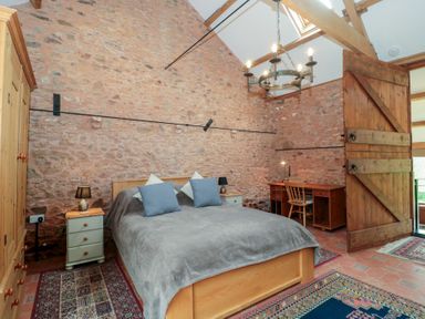 A bedroom with a double bed two pillows bedside tables lamps a wooden wardrobe a desk and chair exposed stone walls and a wooden door at The Barn in Spaxton