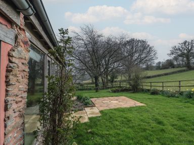 A garden with a stone patio and trees beside a stone and wood building at The Barn in Spaxton