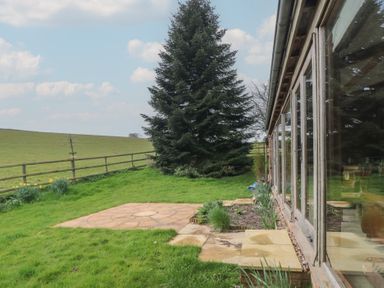 A garden with grass a large evergreen tree and a stone patio next to a building with glass doors at The Barn in Spaxton