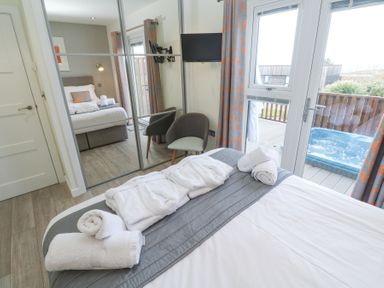 A bedroom with a bed towels and robes a chair mirrored wardrobe a wall mounted tv and door leading to a deck with a hot tub at Hepworth in Portreath
