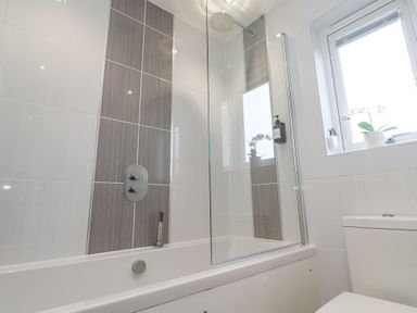 A bathroom with a bathtub shower combination a glass shower screen a window with a potted plant and a toilet at Hepworth in Portreath
