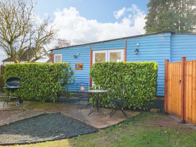 A small patio with a table and two chairs outside a blue wooden building with green hedges at Shed Your Blues Brilley near Kington
