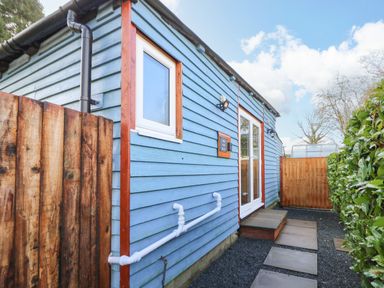 An exterior view of a blue wooden building with a white window and door surrounded by a wooden fence and green hedge at Shed Your Blues Brilley near Kington