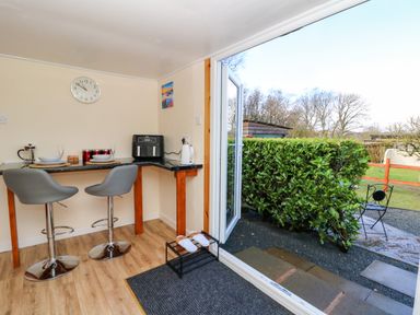 A small dining area with two stools and a countertop with plates and glasses next to an open door leading to a garden with bushes and a metal chair at Shed Your Blues in Brilley near Kington