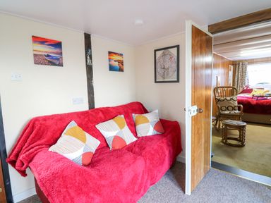 A sitting area with a red covered sofa and pillows next to an open door showing a room with a wooden chair and bed at Shed Your Blues in Brilley near Kington