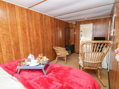 A room with wood panel walls featuring a bed with a red blanket and a breakfast tray a wicker armchair a cushioned wicker chair a black small shelving unit and a TV in the background at Shed Your Blues Brilley near Kington