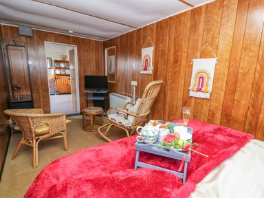 A bedroom with wood panel walls a bed with a red blanket a tray with tea and snacks two rattan chairs a small table a tv and kitchen visible through the doorway at Shed Your Blues in Brilley near Kington