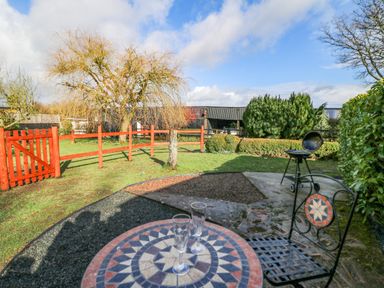 A garden with a red wooden fence a small tree a barbecue and a round mosaic table with two glasses at Shed Your Blues in Brilley near Kington