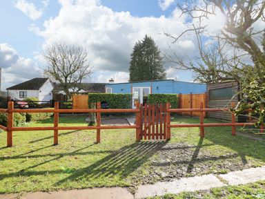A fenced garden with a wooden gate and trees in front of a blue house at Shed Your Blues in Brilley near Kington