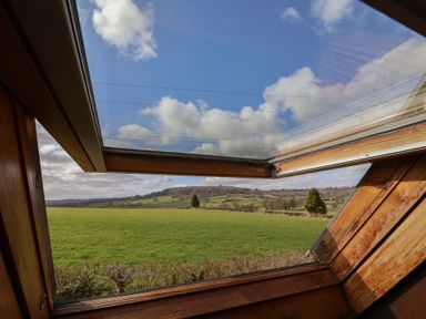 A landscape of green fields and trees seen through an open wooden skylight window at Garden Cottage in Upottery