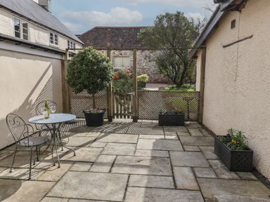 A small patio with a metal table and two chairs a potted tree two rectangular planters and a wooden gate at Garden Cottage in Upottery