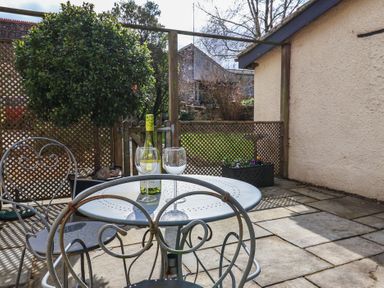 A patio area with a round metal table and two chairs a bottle of wine and two wine glasses a wooden gate and lattice fencing with plants at Garden Cottage in Upottery