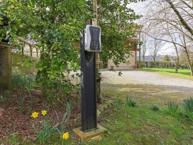 An outdoor electric vehicle charging station on a black post surrounded by plants and trees near a gravel driveway at Garden Cottage in Upottery
