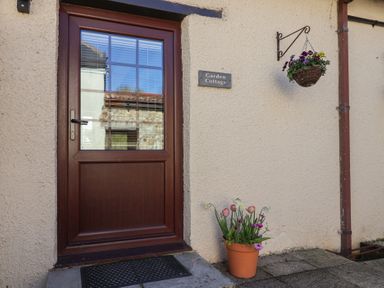 A wooden door with window panes and a hanging basket of flowers with a potted plant below at Garden Cottage in Upottery