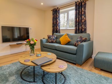 A living room with a teal sofa and matching ottoman a wooden coffee table with flowers and coasters mounted TV and floral curtains at Hideaway in Great Torrington