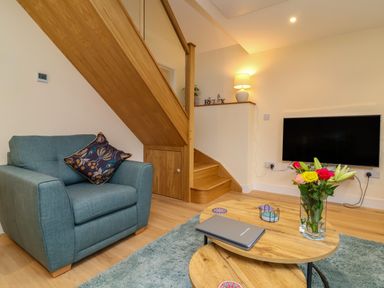 A living room with a blue armchair wooden staircase a wall-mounted television a lamp a wooden coffee table with flowers and a laptop at Hideaway Great Torrington