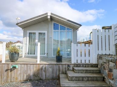 Front view of a small wooden house with glass doors and windows on a raised deck with wooden steps and white fencing Ocean Breeze Abersoch