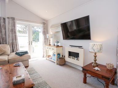 A living room with a beige sofa wooden tables a wall mounted television and glass doors at Ocean Breeze in Abersoch