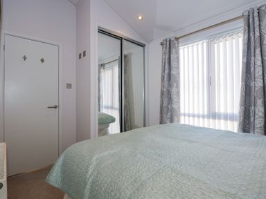 A bedroom with a bed covered by a light blue quilt a window with vertical blinds and patterned curtains a door and a mirrored sliding wardrobe at Ocean Breeze in Abersoch