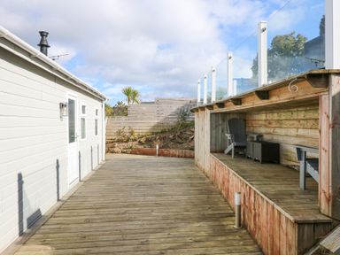 An outdoor wooden deck with seating area and a white building wall with door and windows at Ocean Breeze in Abersoch