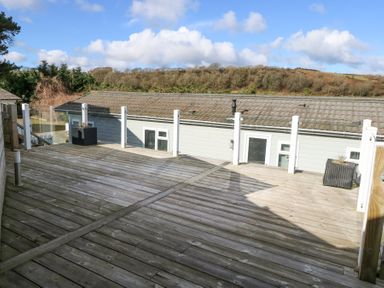 An outdoor wooden deck with glass panel railing and planters overlooking a neighboring building and trees at Ocean Breeze in Abersoch