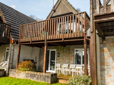 An outdoor patio area with white chairs under a wooden balcony attached to a stone house at Valley Lodge 37 in St Anns Chapel Cornwall