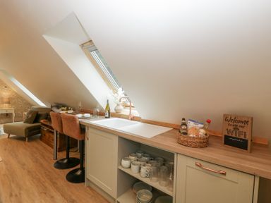 A kitchen area with a sink countertop open shelves with mugs and glasses bar stools and a lounge area at Pheasants Rise Apartment in Ross-On-Wye