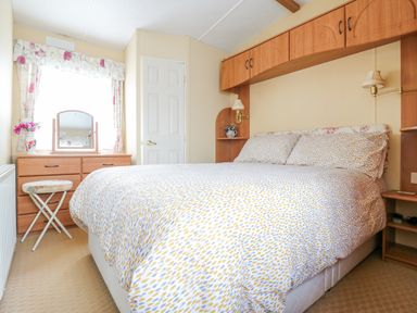 A bedroom with a bed a wooden dresser with a mirror a stool and wooden overhead cabinets at 120 Caravan in Hemsby