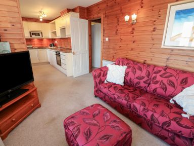 A living room with a red patterned sofa and ottoman a television on a wooden stand and a kitchen with white cabinets at Creag Mhor Lodge Back of Keppoch near Arisaig