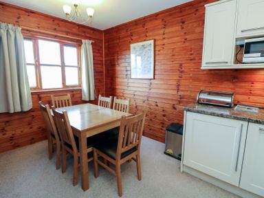 A dining area with a wooden table and six chairs next to a window with curtains in a room with wood-paneled walls at Creag Mhor Lodge Back of Keppoch near Arisaig