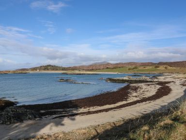 A beach with sand and rocks by a body of water with hills and scattered buildings in the background at Creag Mhor Lodge Back of Keppoch near Arisaig