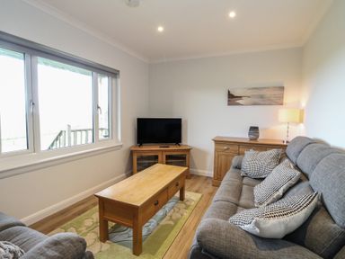 A living room with grey sofas a wooden coffee table a TV on a wooden stand and a sideboard with a lamp at Glen Mhor Lodge in Arisaig