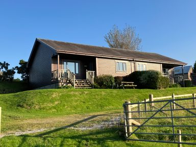A single-story wooden house with two small porches and stairs on a grassy hill with a metal gate in the foreground at Glen Mhor Lodge in Arisaig