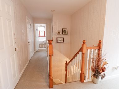 A hallway with beige carpet and wallpaper leading to a bedroom with open doors and wooden staircase banister with a vase of dried flowers at Kerangarth in Corbridge