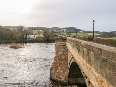 A stone bridge over a flowing river with lampposts and houses in the distance at Kerangarth in Corbridge