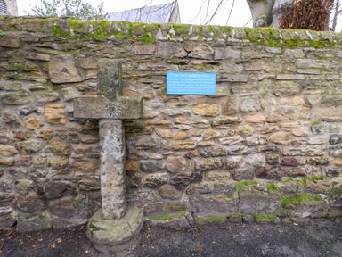 An old stone cross mounted on a stone wall with a blue information plaque attached at Kerangarth in Corbridge
