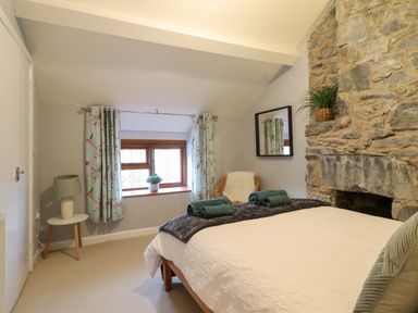 A bedroom with a bed a stone fireplace a window with curtains a small table with a lamp and a chair with a cushion at Gwynfa in Ysbyty Ifan