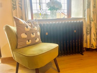 A green chair with a cushion featuring sheep beside a black radiator under a window with floral curtains at Gwynfa in Ysbyty Ifan