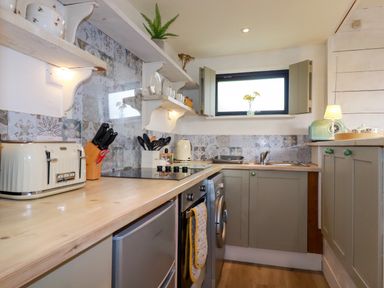 A kitchen with a wooden countertop appliances open window and shelves holding dishes at The Cow Shed in Porthcothan near Mawgan Porth