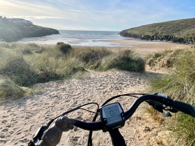 A sandy path with grass on both sides leading to a beach with the sea and hills in the background with bicycle handlebars in the foreground at The Cow Shed in Porthcothan near Mawgan Porth