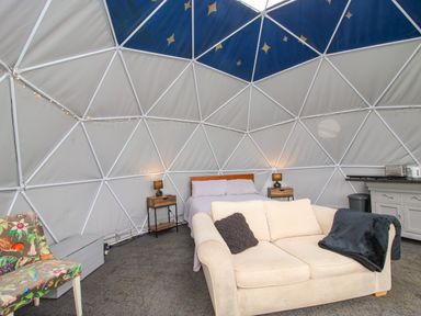 An interior of a dome with a bed two bedside tables with lamps a beige sofa with cushions a floral chair and a kitchenette at Meadow View - Bryn-Mawr Retreat in Brynmawr near Llanymynech