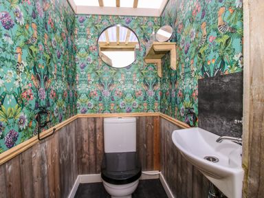 A bathroom with a toilet sink floral patterned wallpaper wooden paneling a round mirror and a small shelf at Meadow View - Bryn-Mawr Retreat Brynmawr near Llanymynech