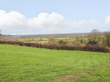 A grassy field with hedges and trees overlooking distant farmland under a partly cloudy sky at Meadow View - Bryn-Mawr Retreat in Brynmawr near Llanymynech