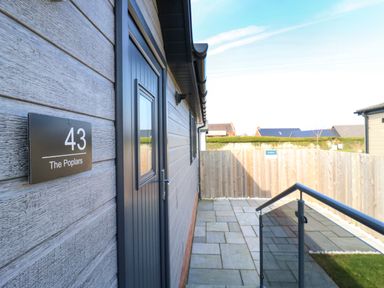 An outdoor patio area with a dark door and a sign reading 43 The Poplars next to a wooden fence and stone tiles at Number 43 The Poplars in Burnham Market