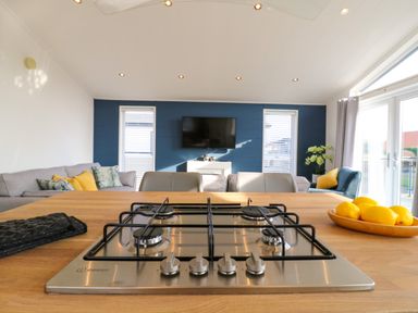 A kitchen island with a gas stove and bowls of lemons overlooking a living room with sofas chairs and a wall mounted tv at Number 43 The Poplars in Burnham Market